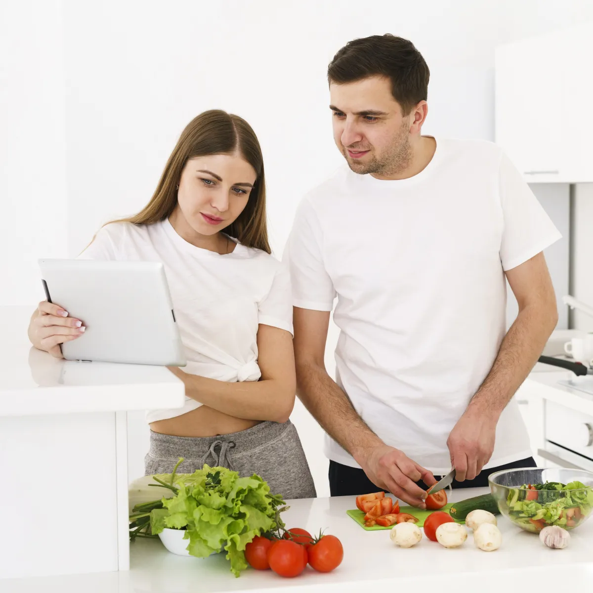 A man and woman in a kitchen preparing a healthy meal together while looking at a tablet, illustrating how men and women may approach the Hyper-Ketosis plan differently based on gender-specific needs and strategies.