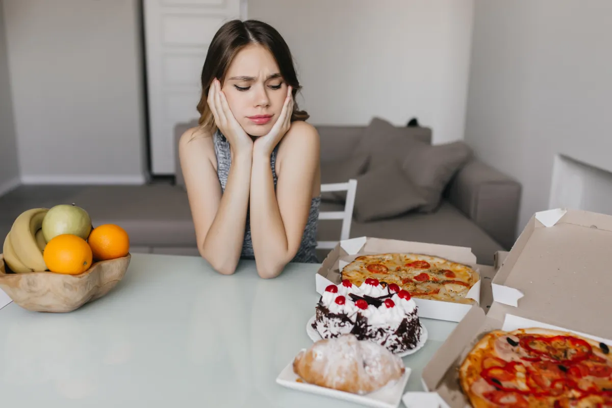 Young woman frowning at pizzas and cakes while a bowl of fruit sits nearby, capturing the struggle with cravings on Hyper-Ketosis