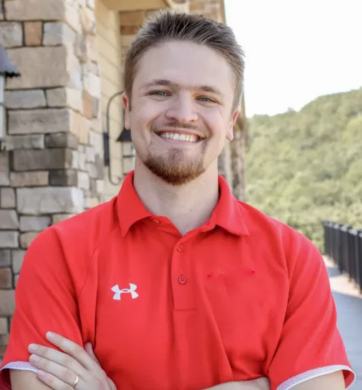 Jackson Lohr stands in front of a stone building with his arms crossed, wearing a red polo shirt and smiling