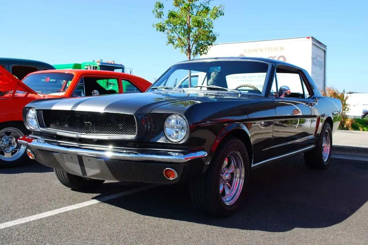 A classic black Mustang car parked in a lot with a red car and a tree in the background, under a clear blue sky.