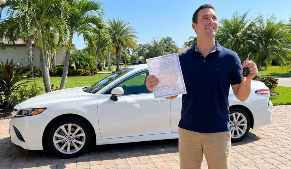 A man holding a vehicle title and car keys in a sunny driveway with palm trees in the background, illustrating how to replace a lost title in Florida.
