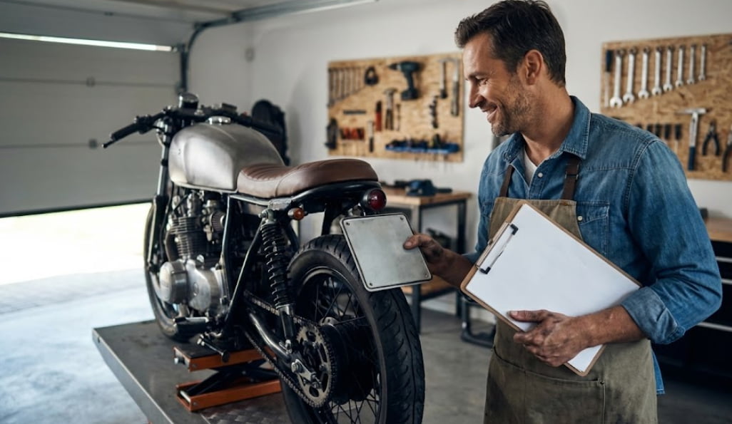 A motorcycle owner in a garage holding a new license plate and registration paperwork for a restored vintage bike, representing successful motorcycle title recovery in the USA.