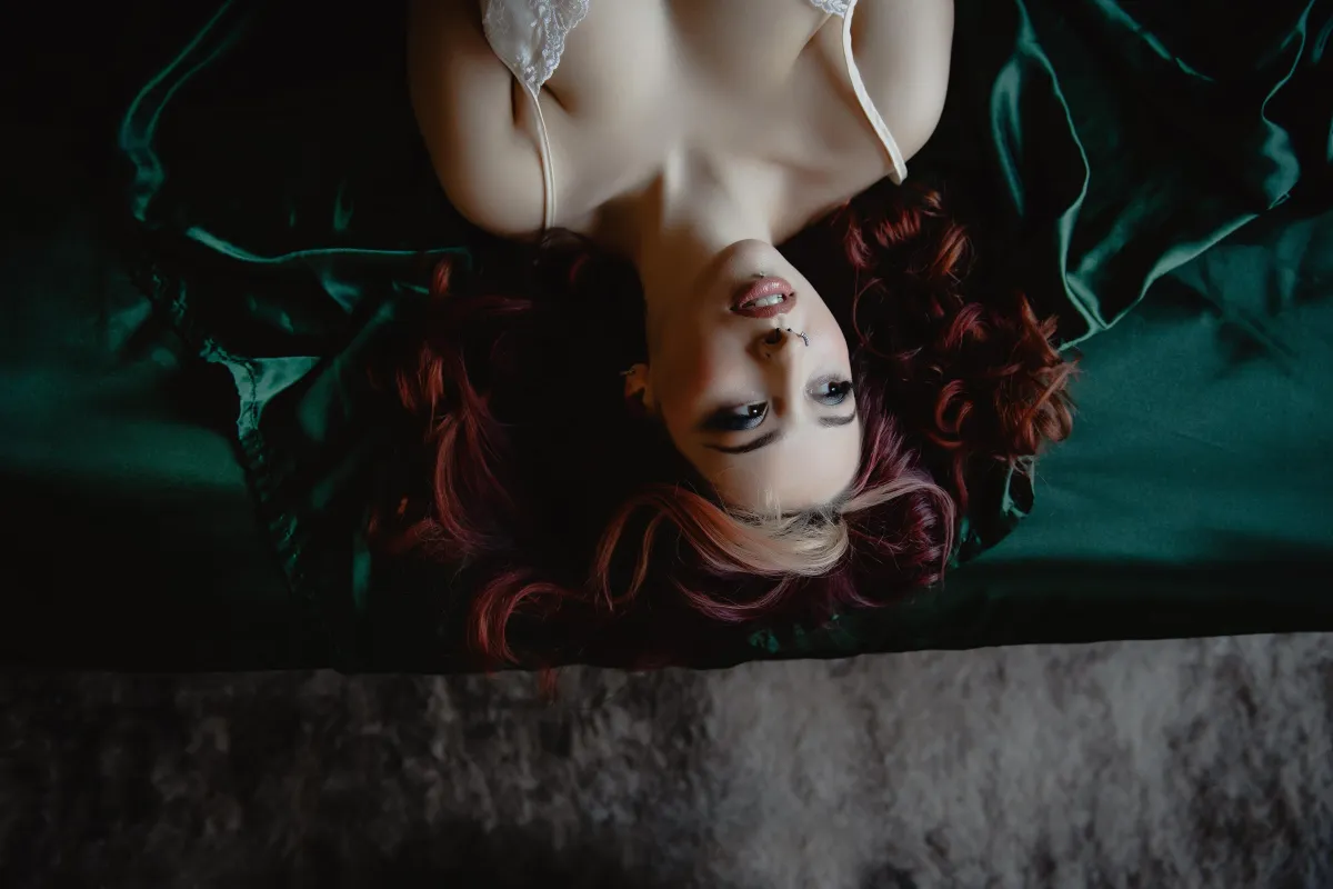 Boudoir photograph of a woman in a satin slip dress posing in a cozy, softly lit bedroom studio