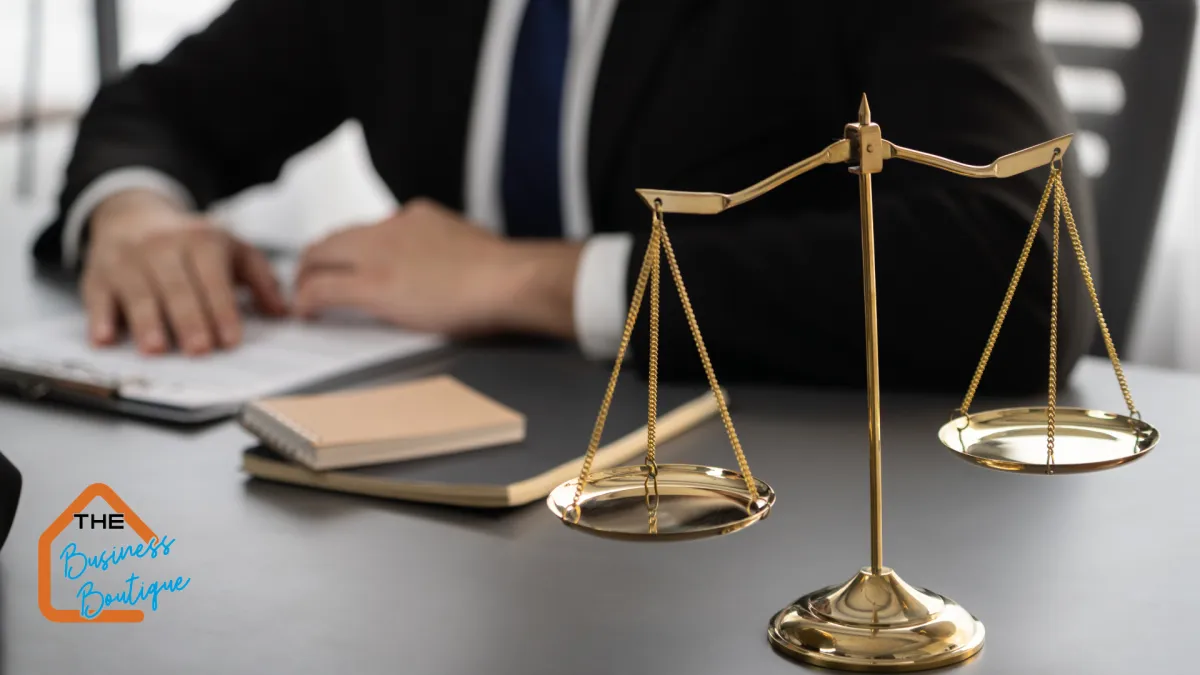 An image of a golden balance scale placed on a desk, symbolizing equilibrium and fairness, with a blurred figure of a professional in business attire working on documents in the background. The Business Boutique logo is displayed in the lower-left corner.