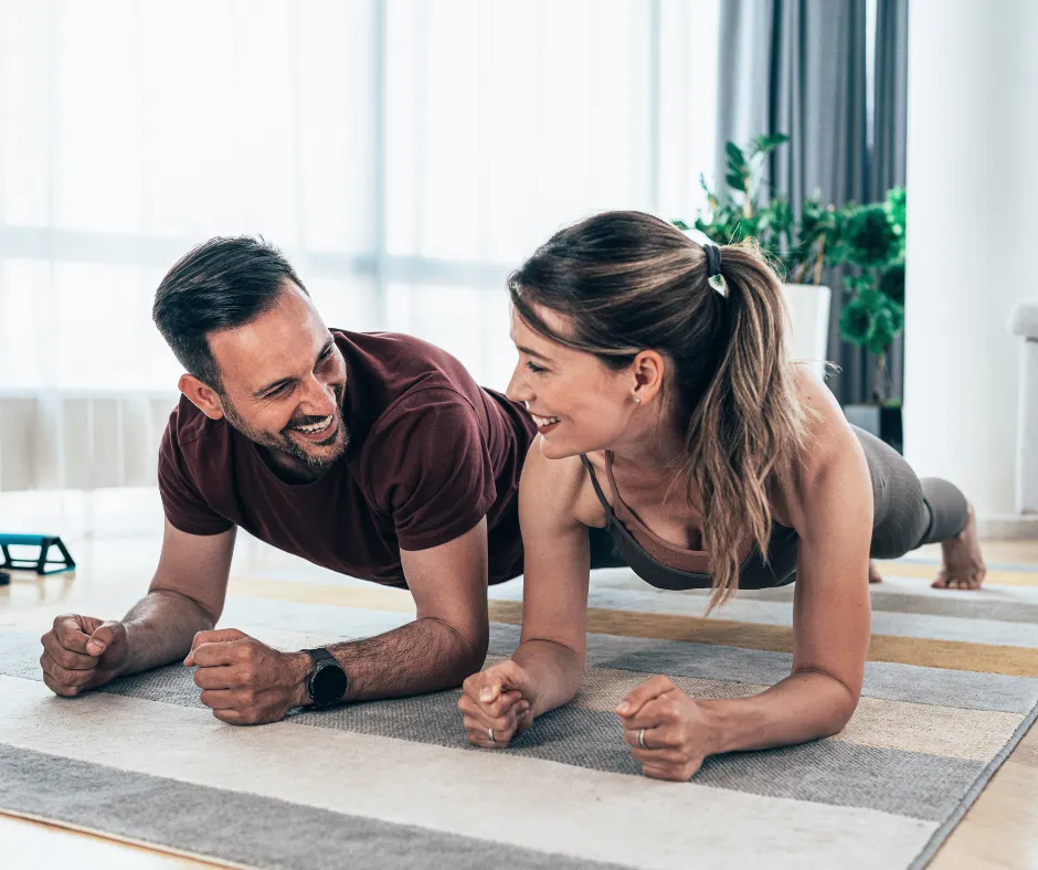 Two young adults holding the plank position and looking at each other smiling.