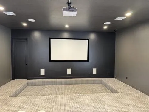 Minimalist home theater room with projector, wall-mounted screen, built-in speakers, and tiered carpeted seating platform.