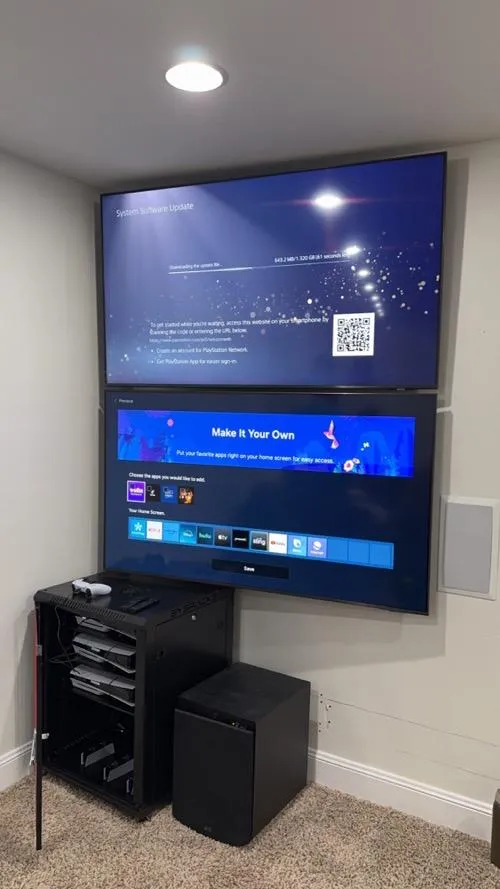 Home entertainment system with dual wall-mounted TVs, gaming rack with consoles, and subwoofer on carpeted floor.