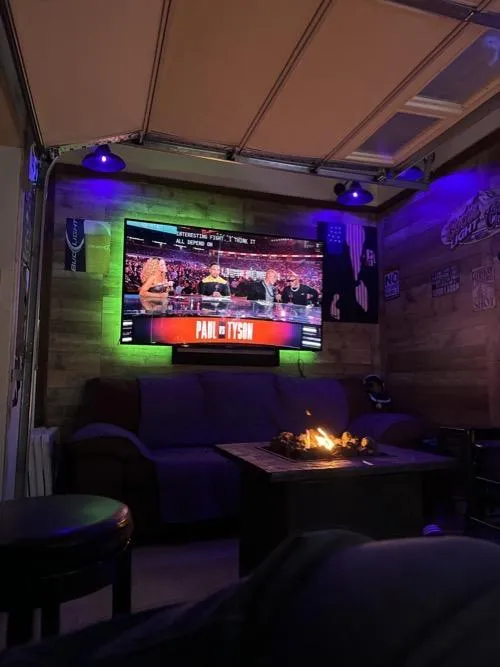 Cozy home theater setup with wall-mounted TV showing a Paul vs Tyson fight, purple and green LED lights, couch seating, and a fire pit table.