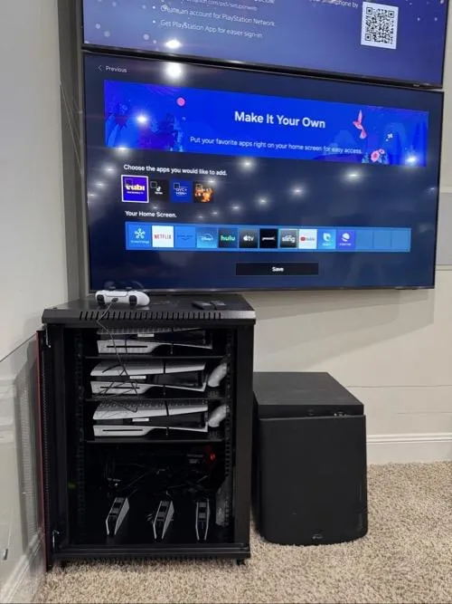 Home entertainment setup with wall-mounted TV, streaming apps on screen, gaming consoles stacked in a rack cabinet, and subwoofer beside it.