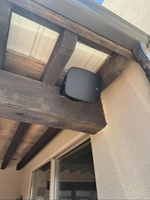 Outdoor mounted black speaker installed under wooden pergola beams near a house wall.