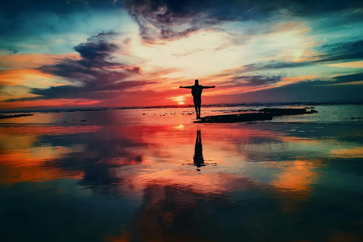 Person standing with open arms at the edge of the water during a vibrant sunset, symbolizing peace, spirituality, and connection with nature.
