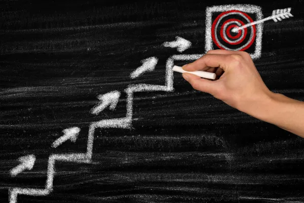 Hand drawing chalk arrows up a staircase toward a target on a blackboard