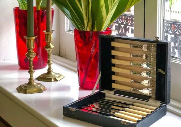 Vintage ivory-handled cutlery set displayed in an open case on a windowsill with brass candlesticks, red glass vases, and green foliage in the background at Fireside Antiques in Bowie, Maryland