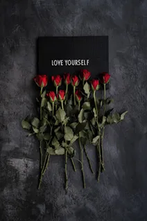 A flat-lay arrangement of red roses placed beneath a black letter board that reads “LOVE YOURSELF,” set against a dark, textured background.