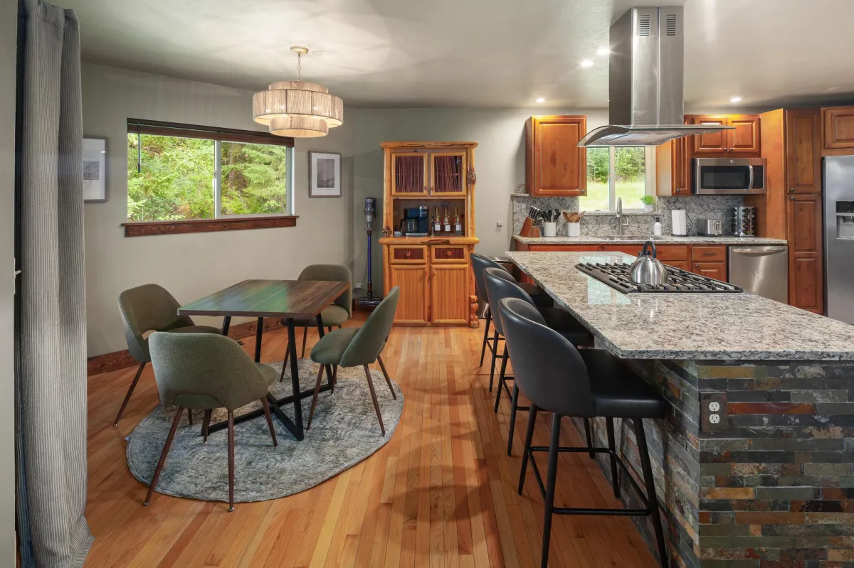 Kitchen with Dining Table & Island Seating – Bright kitchen with granite island seating and dining table, perfect for group meals. Fully stocked for cooking, with views of the surrounding forest near Glacier National Park.