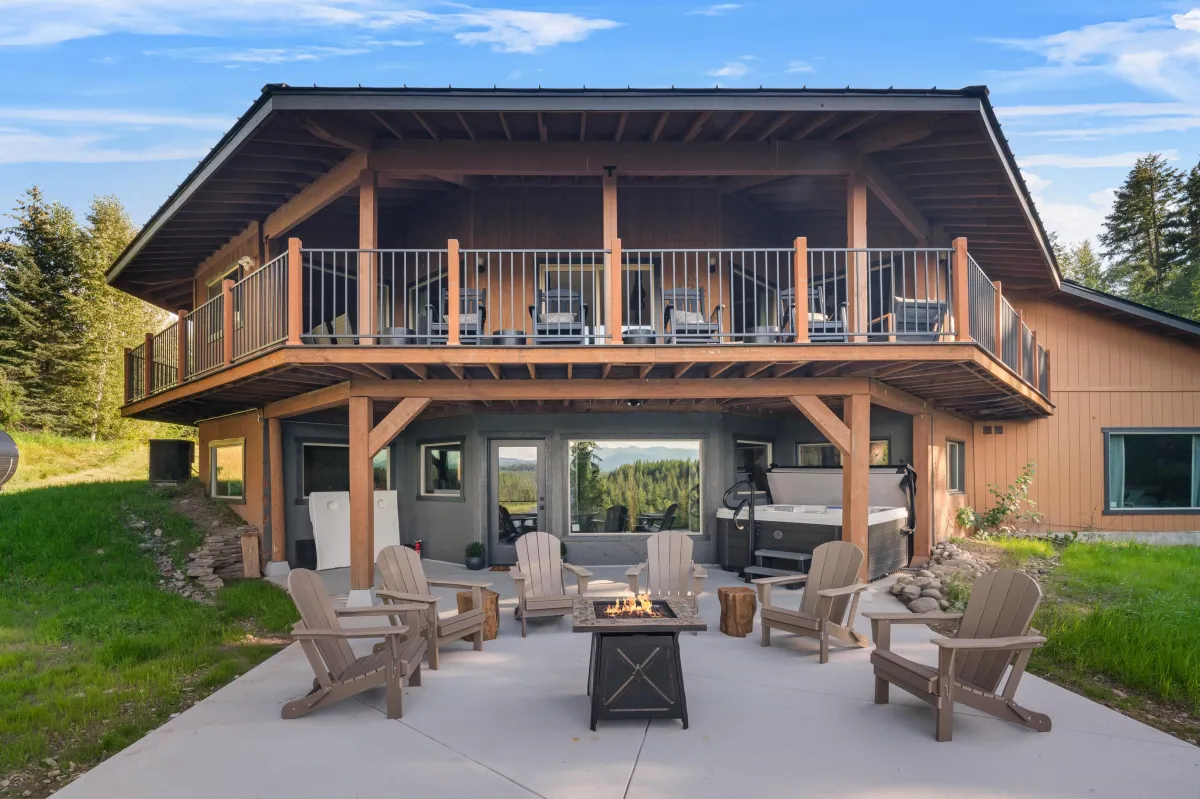 View of the lodge from the river side, showcasing the upper deck, covered lower patio, and fire pit table area. Surrounded by nature in a private 3-acre Montana setting near Glacier National Park.