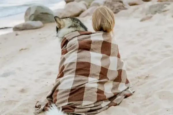 A woman and her dog on a beach wrapped in a blanket