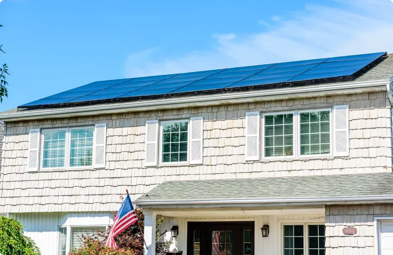 A charming Mapleton, UT home with a modern solar panel installation on the roof, featuring light-colored siding, multiple windows with shutters, and a welcoming front porch adorned with an American flag, set against a clear blue sky.