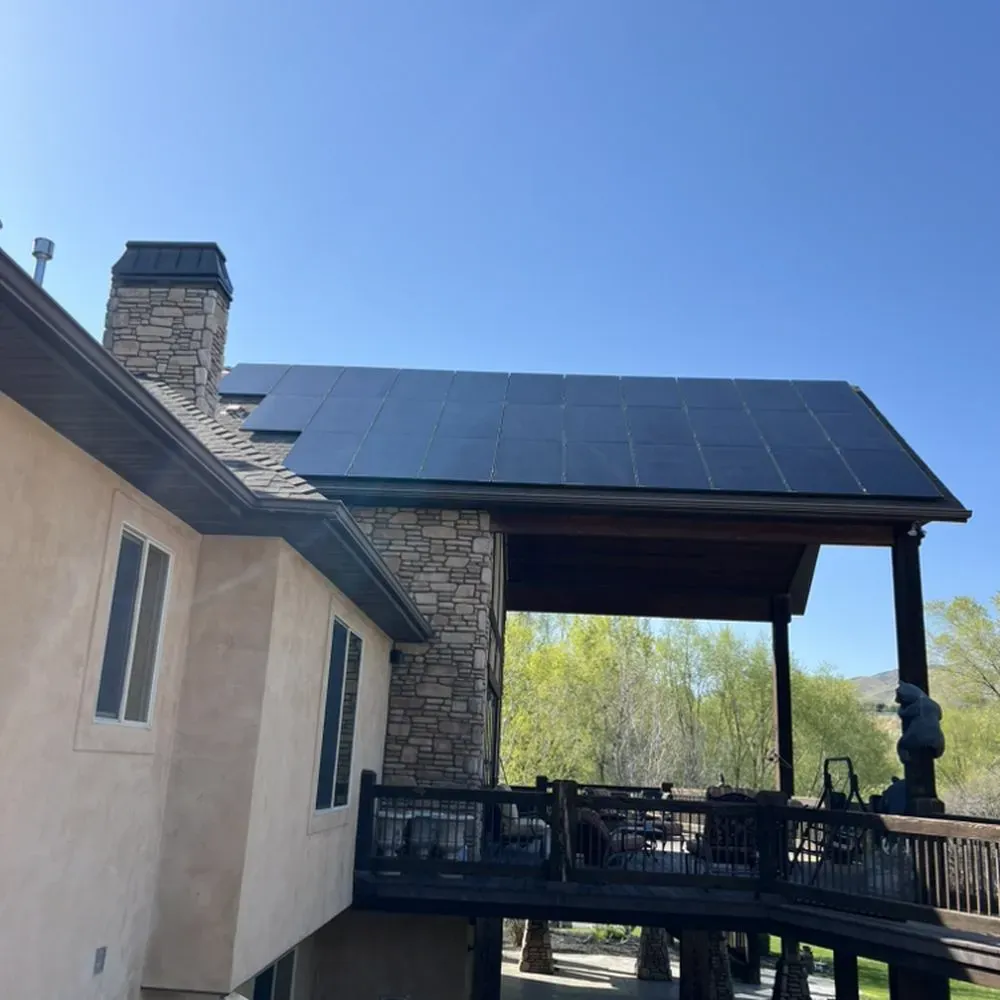 A modern Benjamin, UT home featuring a robust solar panel array on the roof, complemented by a stone chimney and stucco exterior, with a spacious wooden deck and outdoor seating area, set against a backdrop of lush greenery and a clear blue sky.
