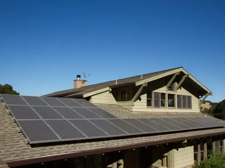 A picturesque Palmyra, UT home with a sleek solar panel array covering the roof, showcasing a classic design with light green siding, multiple windows with shutters, and a prominent chimney, all under a vibrant blue sky with surrounding greenery.