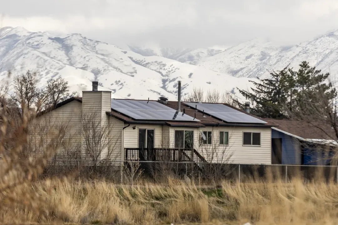 A cozy Spring Lake, UT home with a solar panel array on the roof, featuring light siding, a wooden deck with stairs, and a fenced yard, set against a backdrop of snow-capped mountains and golden wild grasses under a cloudy sky.