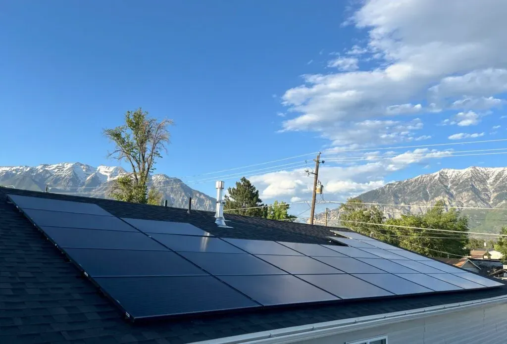 A modern Springville, UT home with an expansive solar panel array on the roof, surrounded by lush trees and set against a stunning backdrop of snow-capped mountains under a partly cloudy blue sky, with power lines stretching across the horizon.