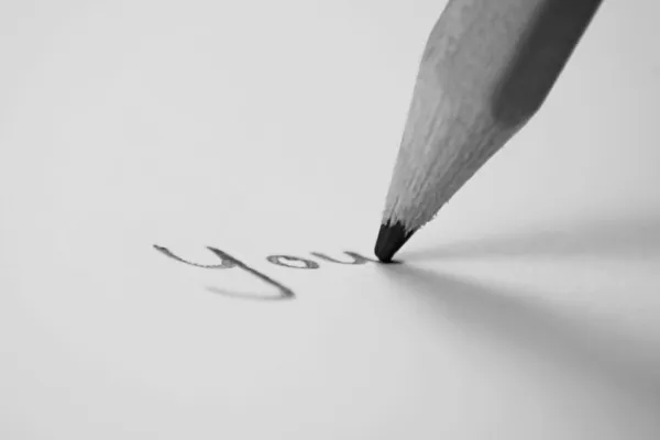 Grayscale image of the tip of a pencil writing the word you on a white sheet of paper.