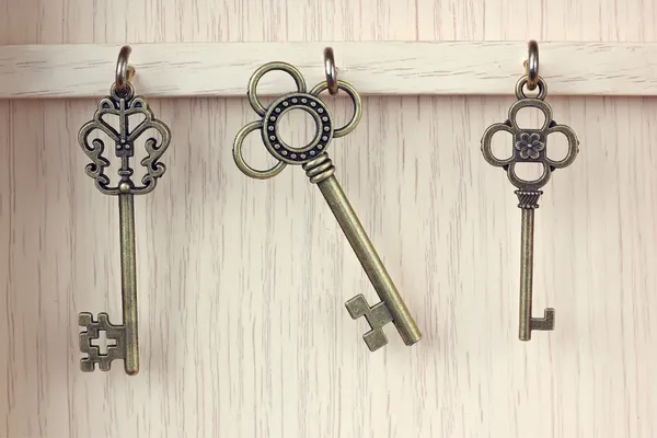 Three old, brass keys hanging from hooks on a light brown wooden wall.