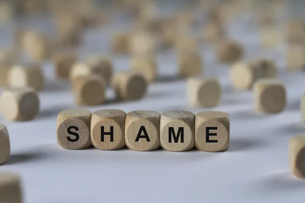 A group of wooden letter cubes like from a game with random letters in the background and the word shame spelled out with cubes in the foreground.