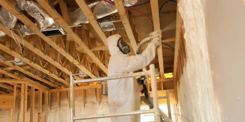 Technician in protective suit applying spray foam insulation to interior walls of a residential building for energy efficiency and air sealing.