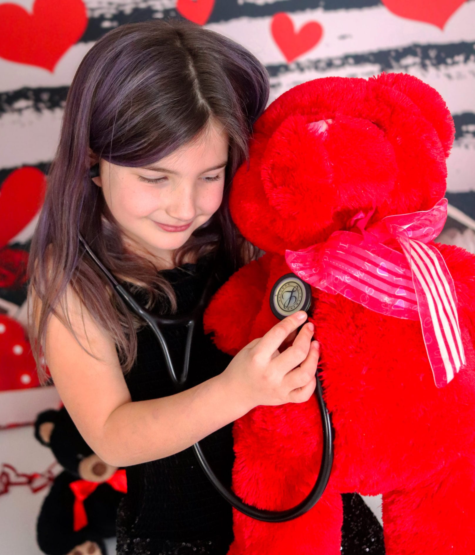 Smiling young girl using a stethoscope to listen to the chest of a large red teddy bear against a Valentine's heart backdrop