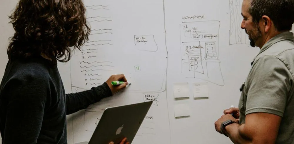 A woman holding a laptop speaks to a man while they stand in front of a whiteboard covered with notes