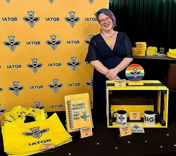 Lisa standing behind a display table with various yellow and black branded items featuring the "IATQB" logo.