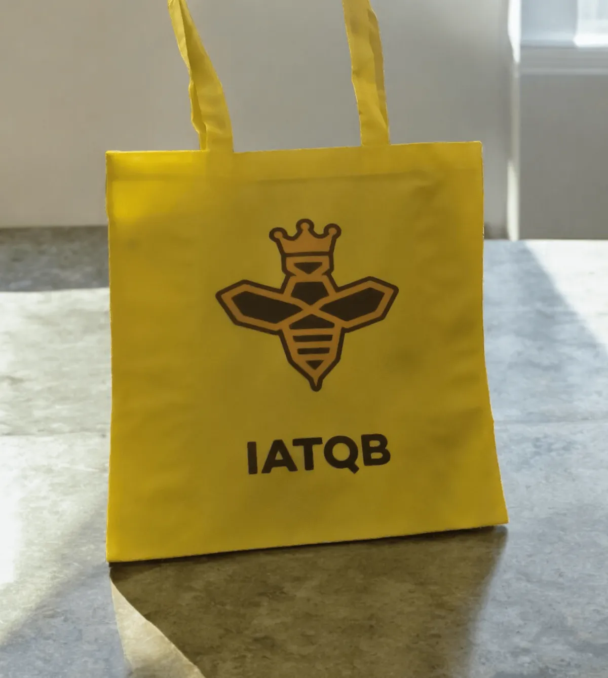 A yellow tote bag with a black and yellow bee on with the letters IATQB underneath
