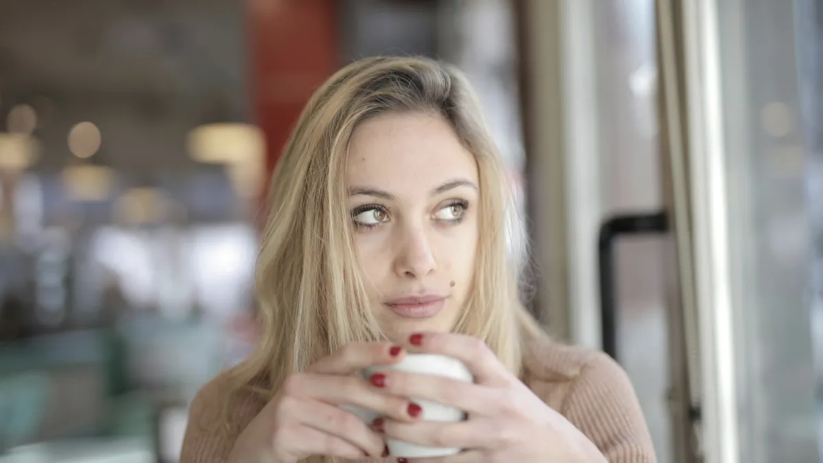 A thoughtful woman holding a white mug, sitting by a window with soft natural light, wearing a cozy sweater, reflecting on healing, self-love, and personal growth during the holidays.