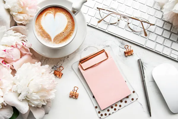 desk filled with coffee, keyboard, flowers, glasses and notepad and pen