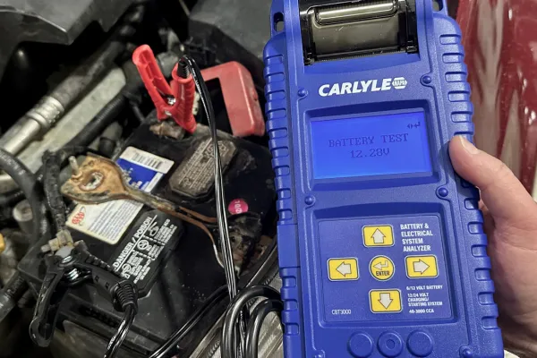 Technician holding a hand-held battery tester showing that vehicle battery has 12 volts