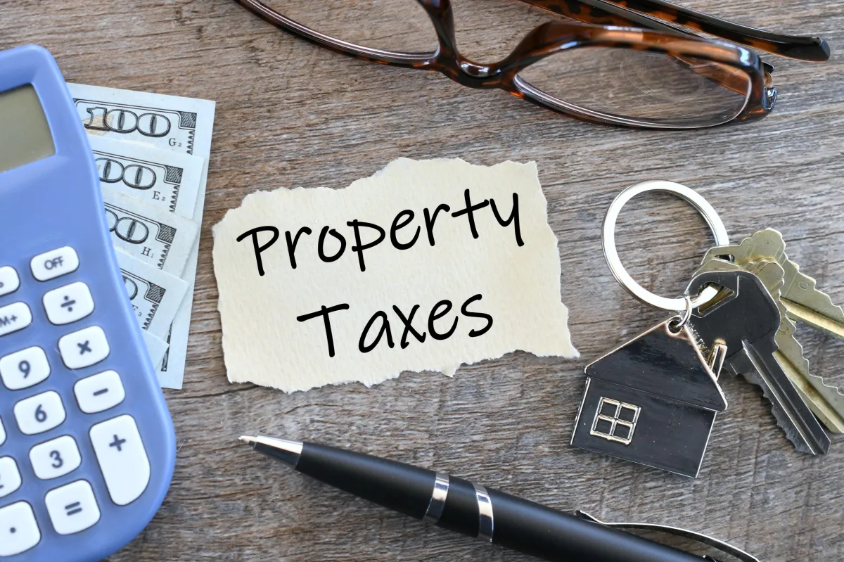 Paper that says Property Taxes with house keys calculator cash money reading glasses and ink pen flat lay.