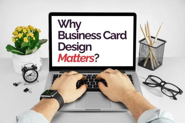 A laptop on a clean, white workspace displays the question ‘Why Business Card Design Matters?’ in bold text. A pair of hands hovers over the keyboard, suggesting active work or research. On the left, a potted plant with yellow flowers and a small black alarm clock add a touch of color and functionality. On the right, a pencil holder and eyeglasses rest on the desk, emphasizing a professional setting. This scene highlights the importance of high-quality, thoughtful business card design in a modern, organized workspace.