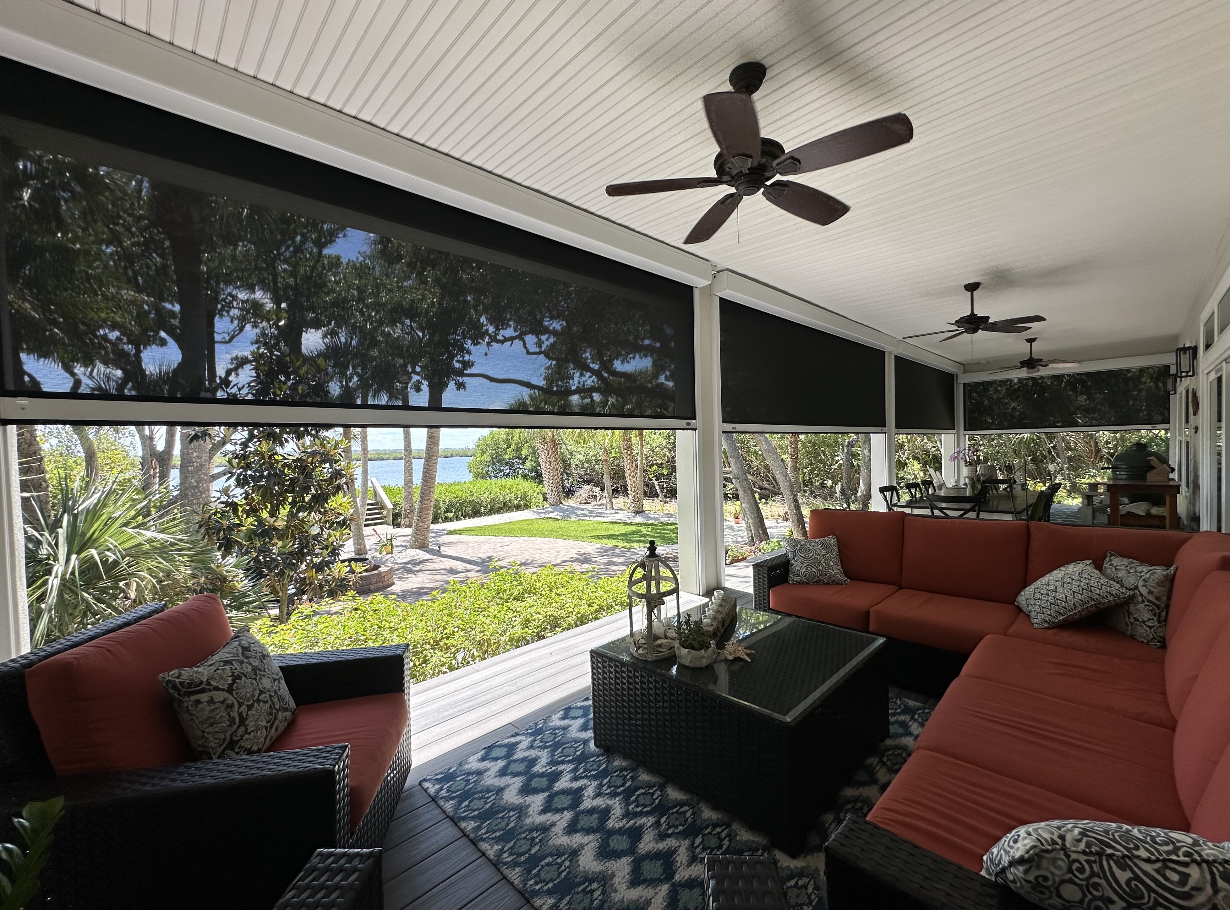 A serene screened-in porch with hurricane protections and a waterfront view, featuring cozy seating with red cushions, ceiling fans, and a patterned blue rug, surrounded by lush greenery outside.