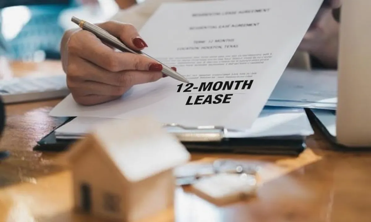 A close-up shot of a person with red nail polish holding a silver pen, poised to sign a "RESIDENTIAL LEASE AGREEMENT" document which is clearly visible. The document specifies "TERM: 12 MONTHS" and "LOCATION: HOUSTON, TEXAS". In the foreground, slightly out of focus, sits a small wooden model house. The overall scene suggests the process of finalizing a 12-month lease agreement in Houston.