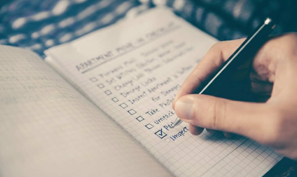Close-up photo of a person's hand using a pen to check the box next to 'Pack' on a handwritten list titled 'Apartment Move-In Checklist' in a spiral notebook.