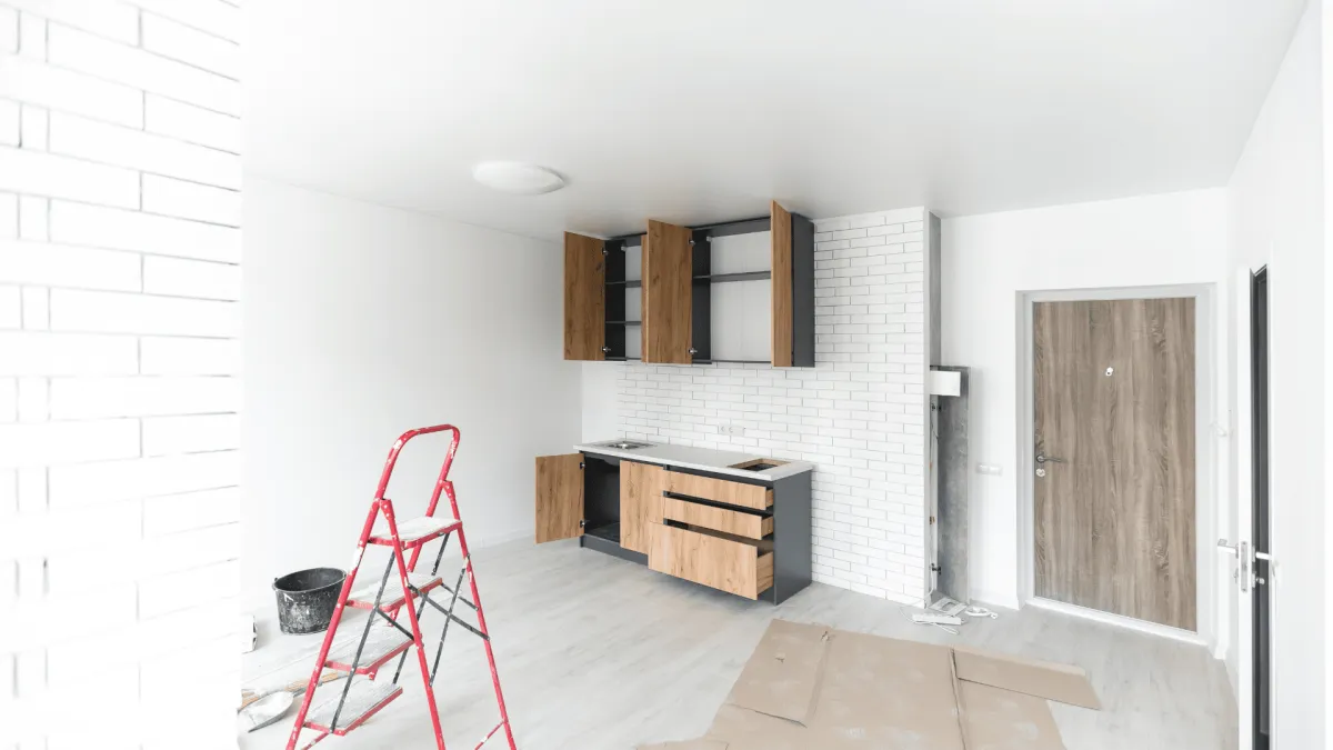 Installation de cuisine moderne avec meubles en bois gris portes ouvertes, carreau blanc mur, échelle rouge au sol en chantier.