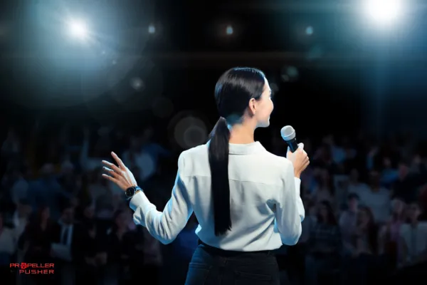 Woman standing on stage with spotlight, audience in the background.