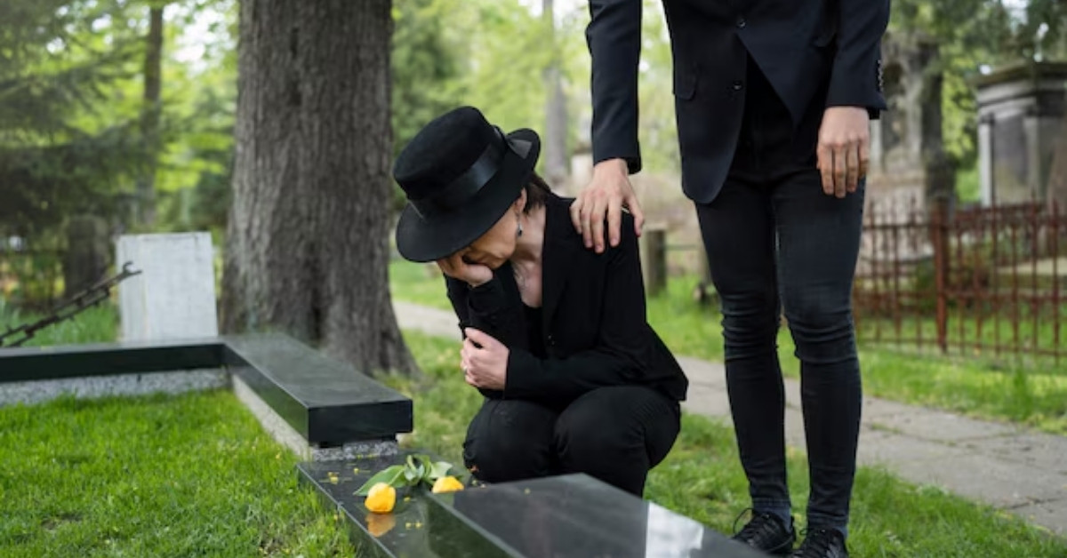A grieving person dressed in black kneels beside a grave while another person offers comfort with a hand on their shoulder in a cemetery.