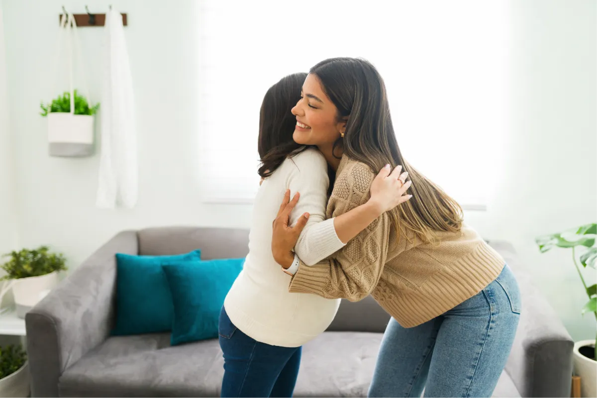 Two women share a heartfelt hug by a couch, highlighting support for postpartum emotional challenges