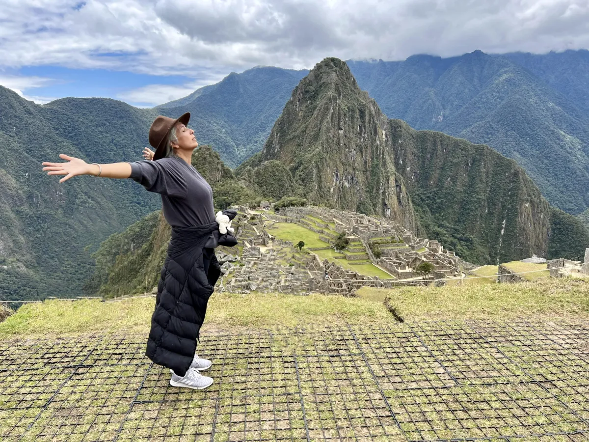 Woman standing triumphantly at Machu Picchu overlook, arms outstretched embracing expansive Andes mountains under dramatic clouds, embodying awakening and embodied sovereignty