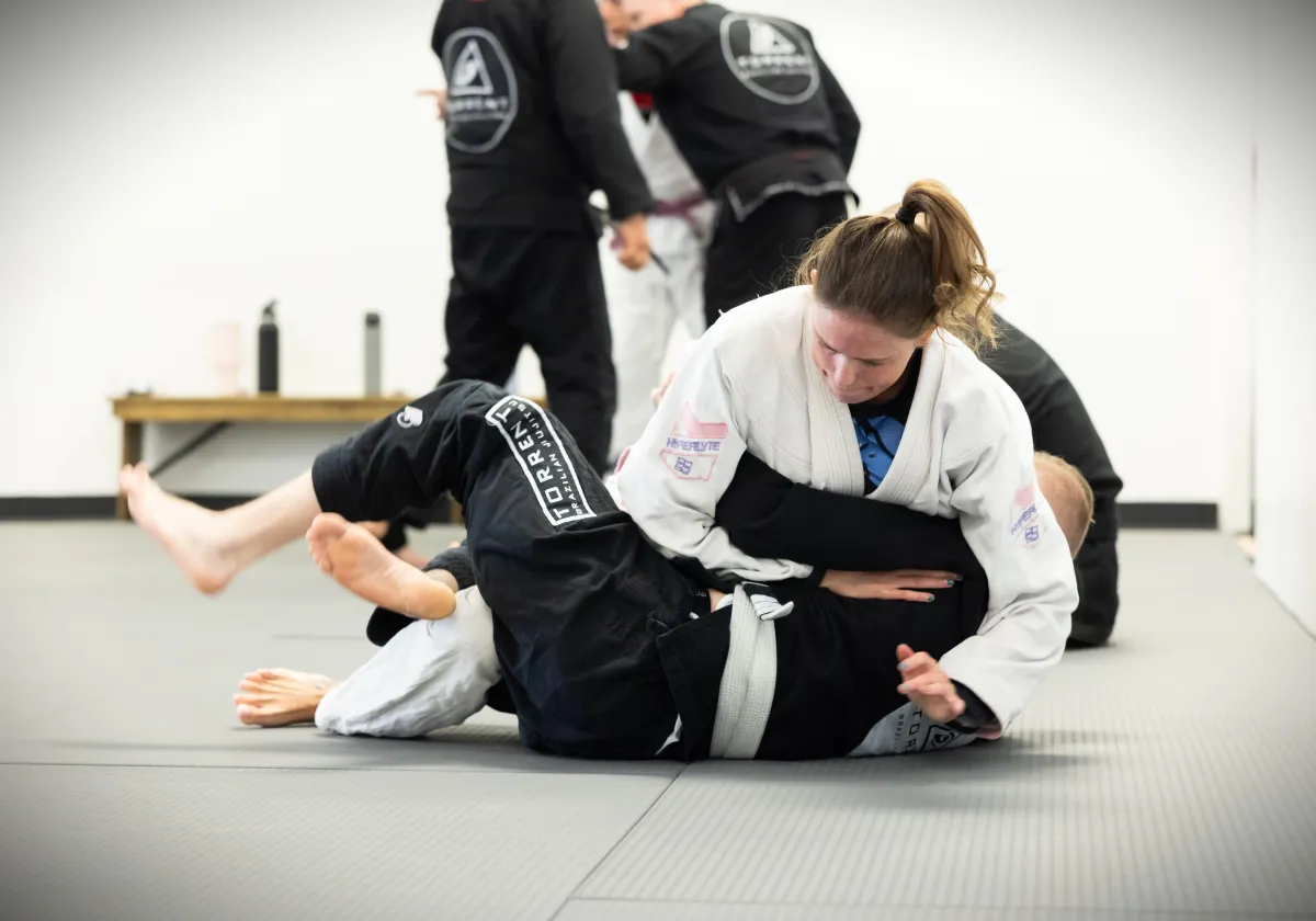 A woman in a gi practicing grappling techniques during a Brazilian Jiu-Jitsu class, focused and in control on the mat.