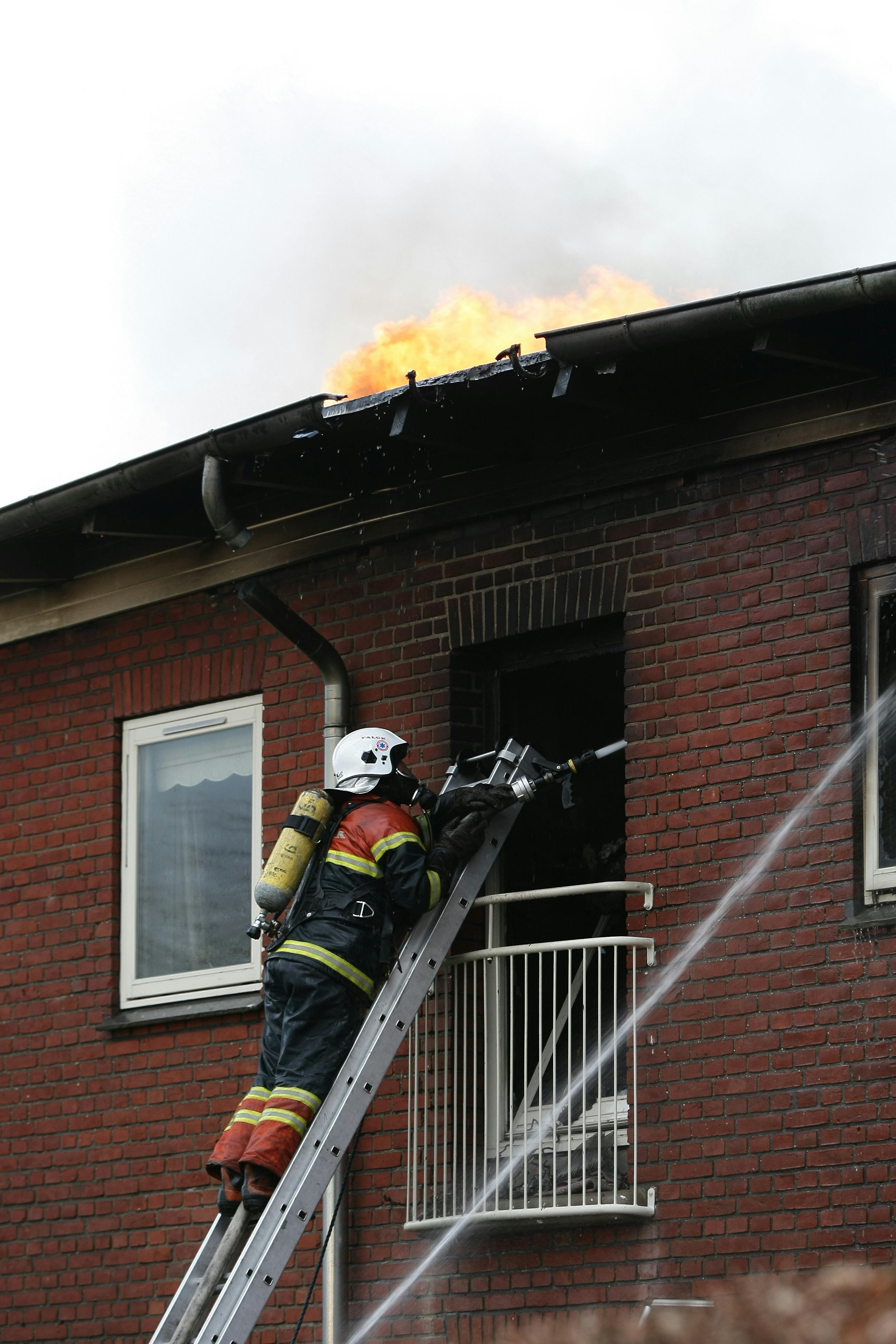 firefighter on ladder reaching into a building on fire with a hose