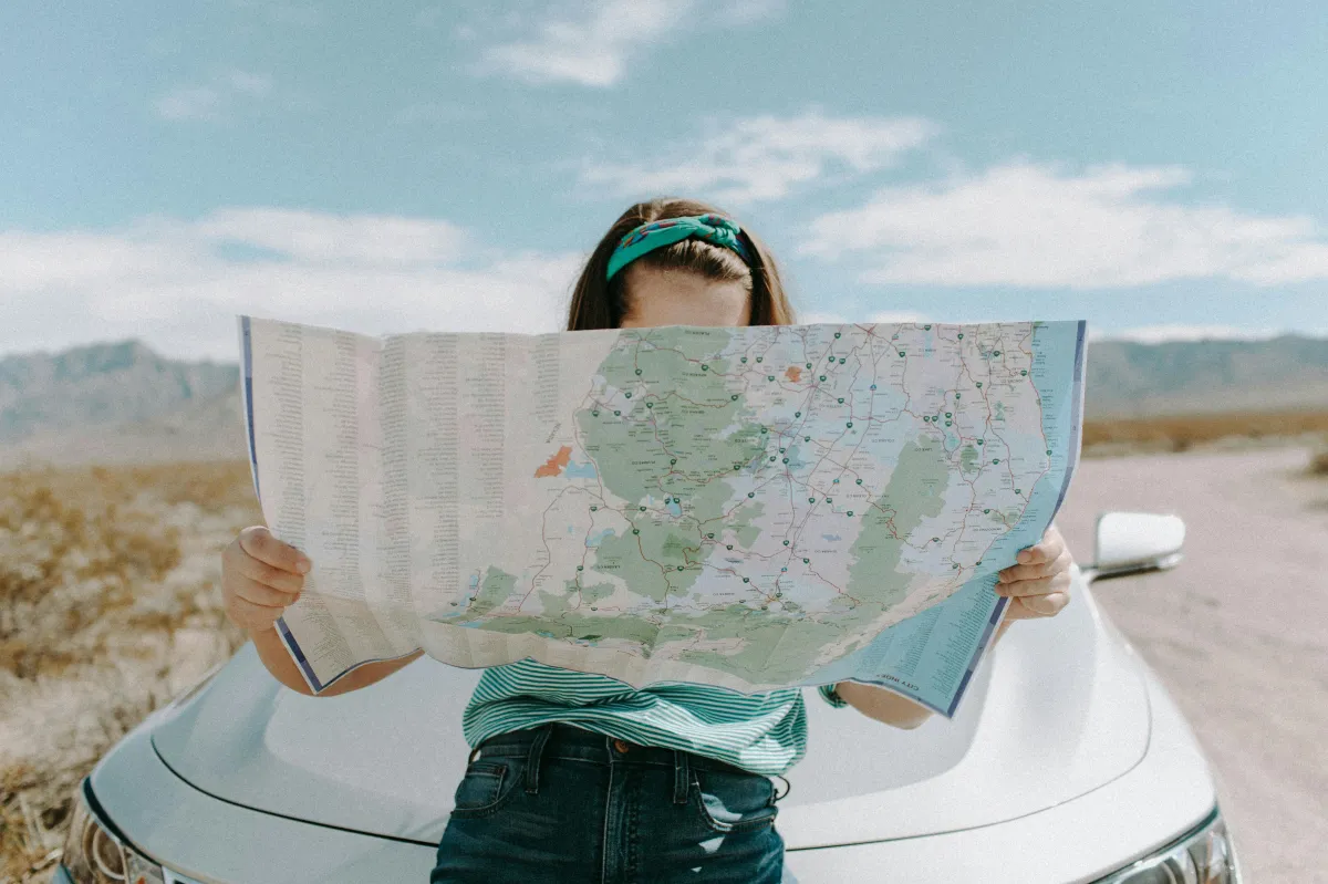 A woman standing in the desert leaning on a car, holding a large map that covers her face. The background shows mountains and a blue sky, symbolizing travel, curiosity, and mental exploration.
